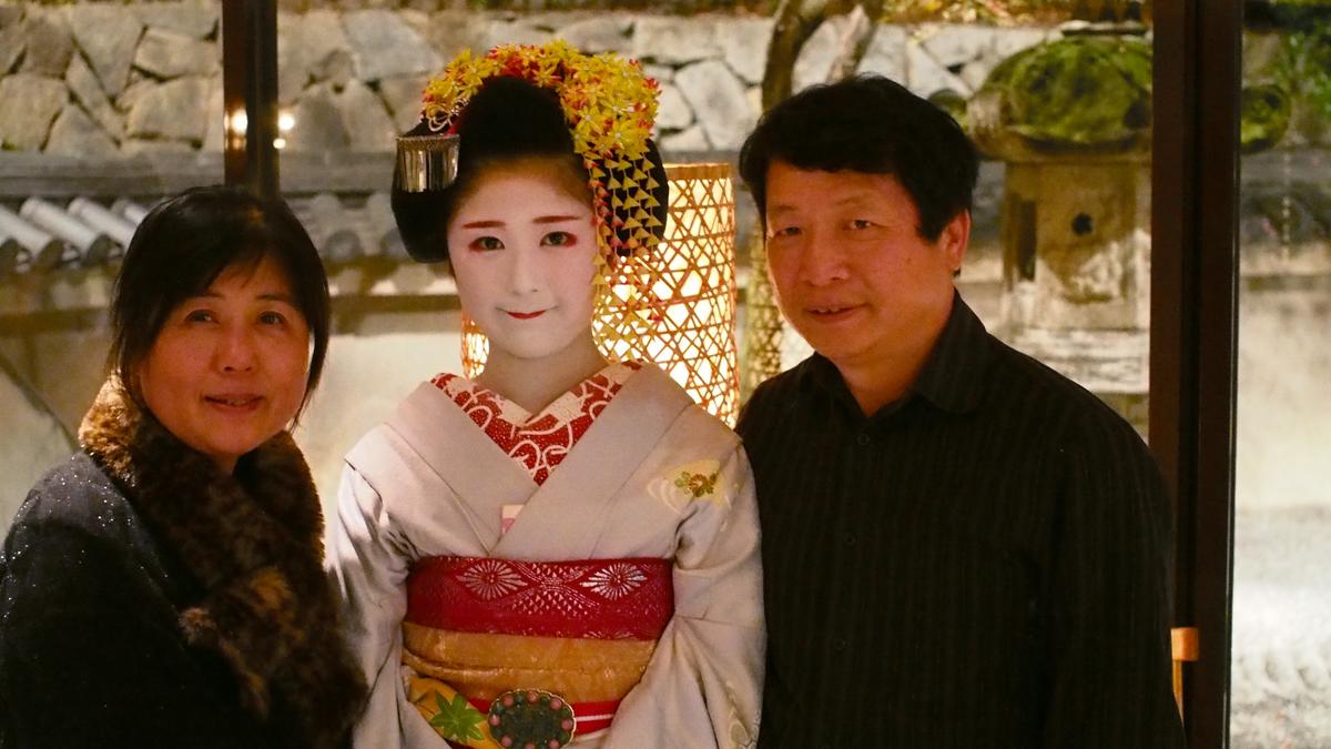 A Maiko in traditional white makeup and kimono smiles between a man and a woman.