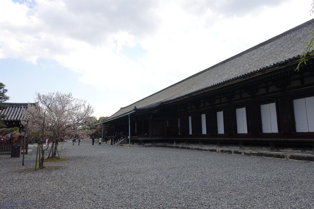 A long, dark traditional Japanese building with a tiled roof stands beside a gravel courtyard with a blooming cherry tree.