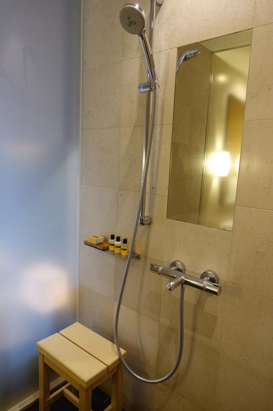 A modern shower with beige tiles, a handheld shower, a wooden stool, and a recessed mirror with bath amenities.