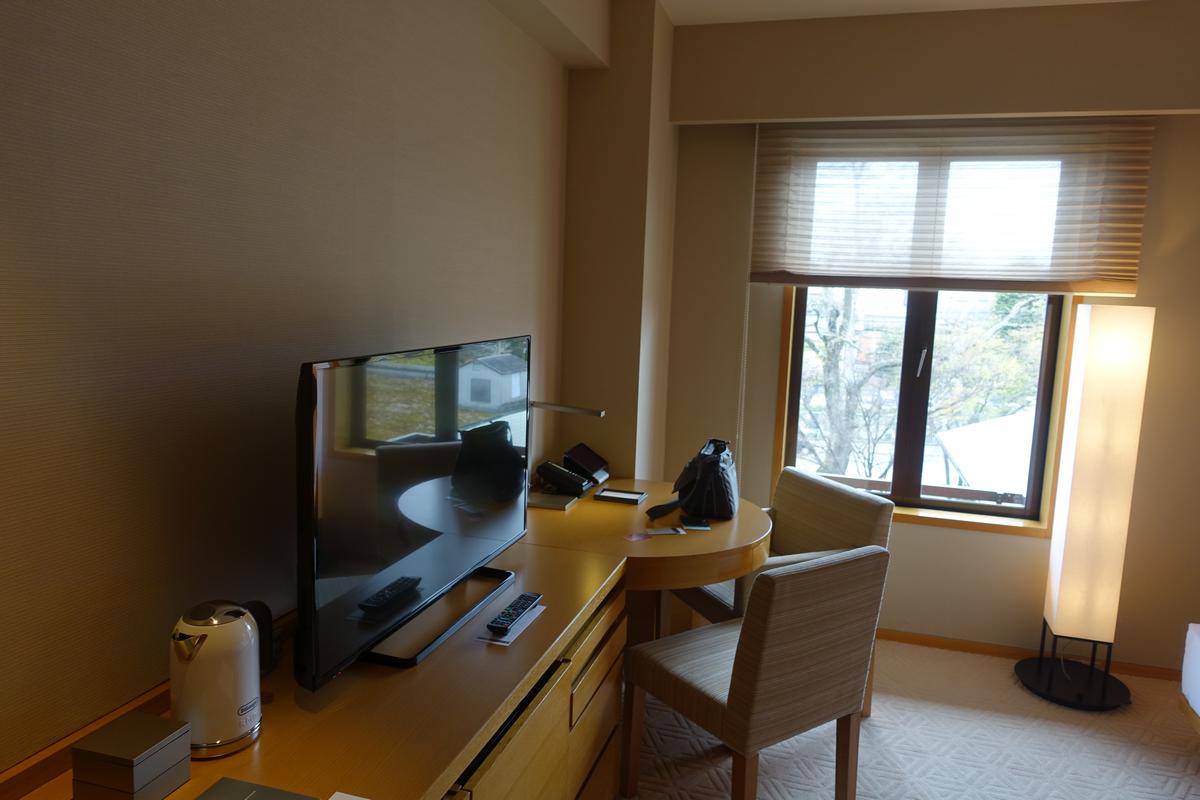 A hotel room featuring a flat-screen TV, a small table with chairs, and a window with blinds overlooking trees.
