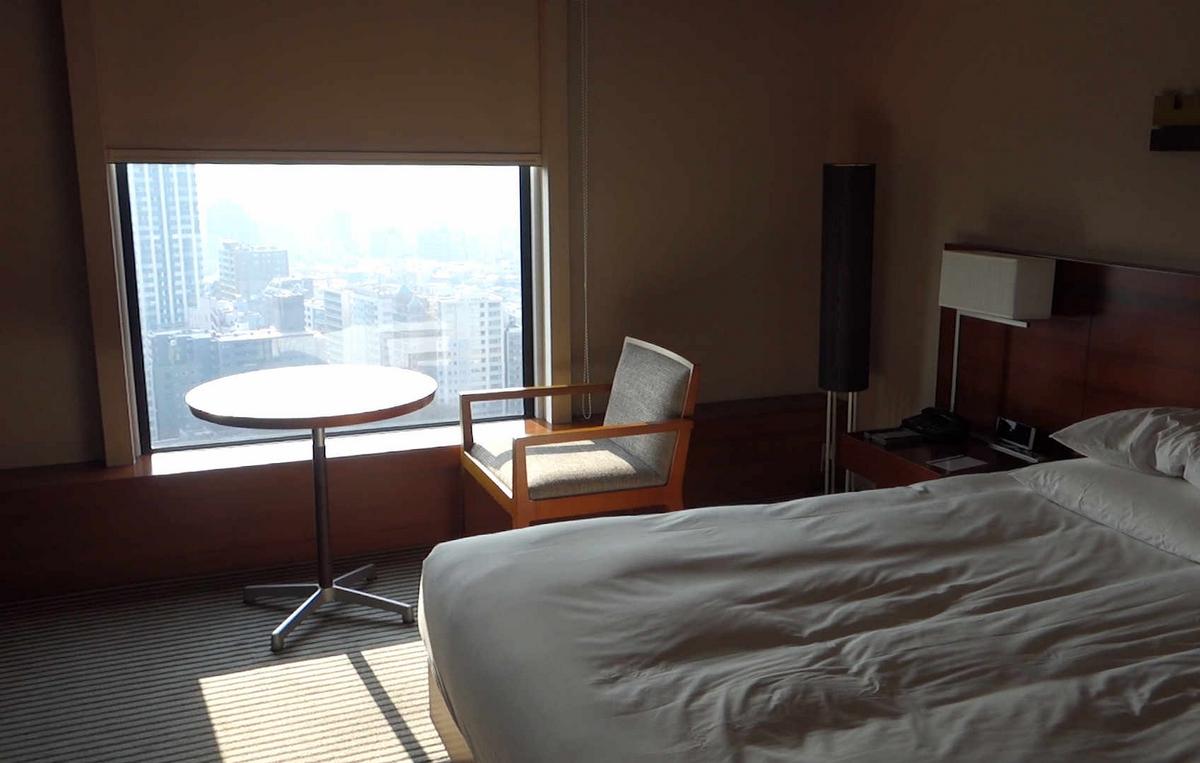 A hotel room with a bed, a table and chair by a window with a city view.