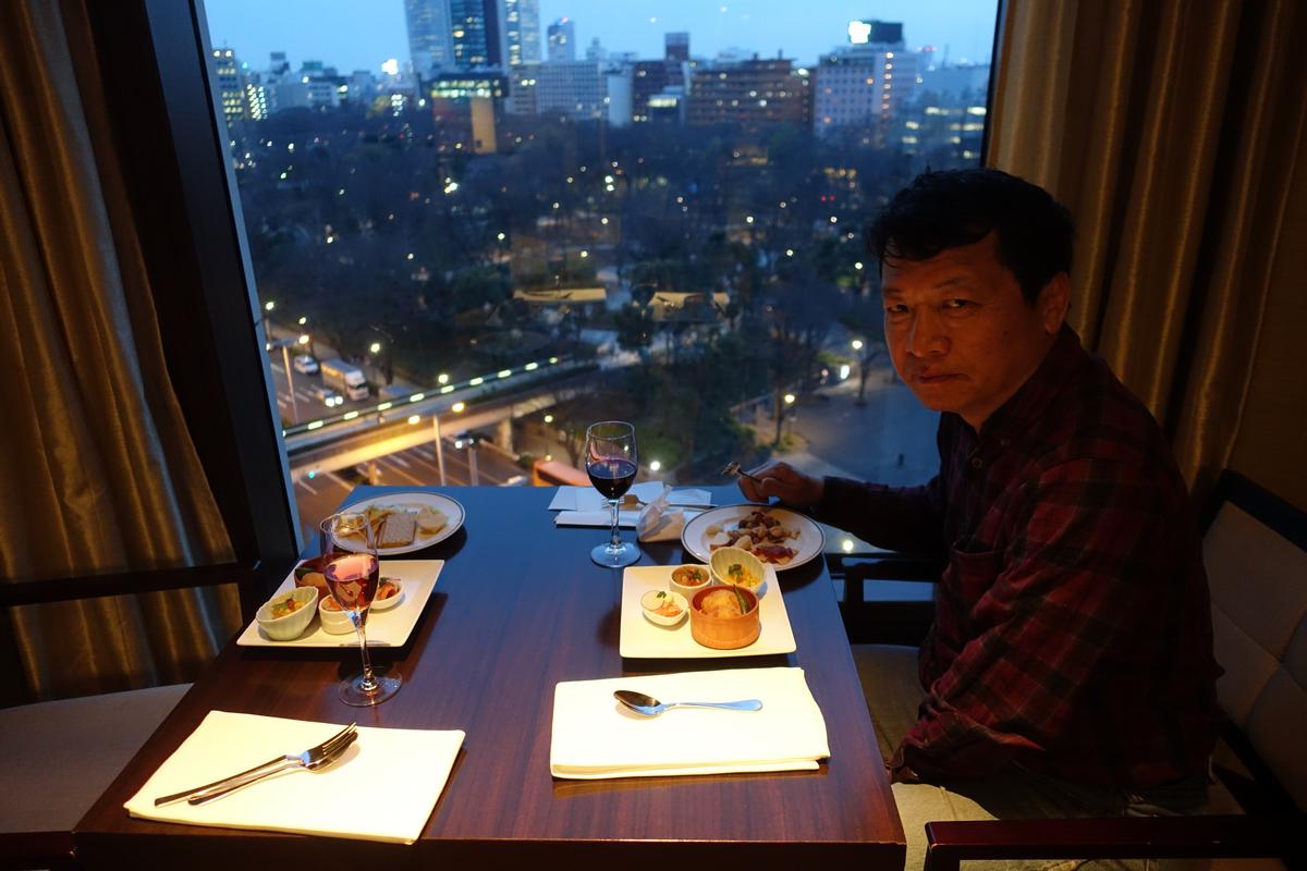 A man looks at the camera while dining at a table by a window with a city view at dusk.