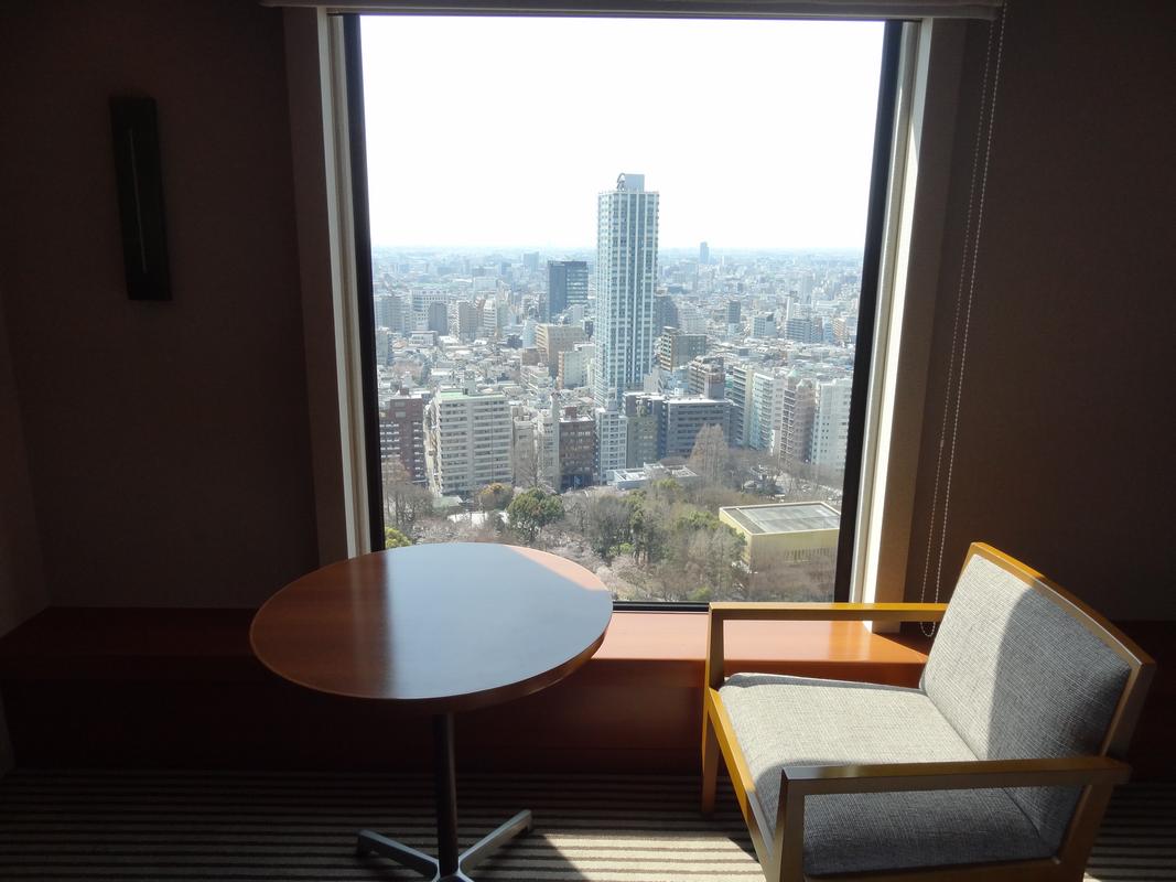 A hotel room with a table and armchair next to a large window overlooking the Tokyo cityscape.