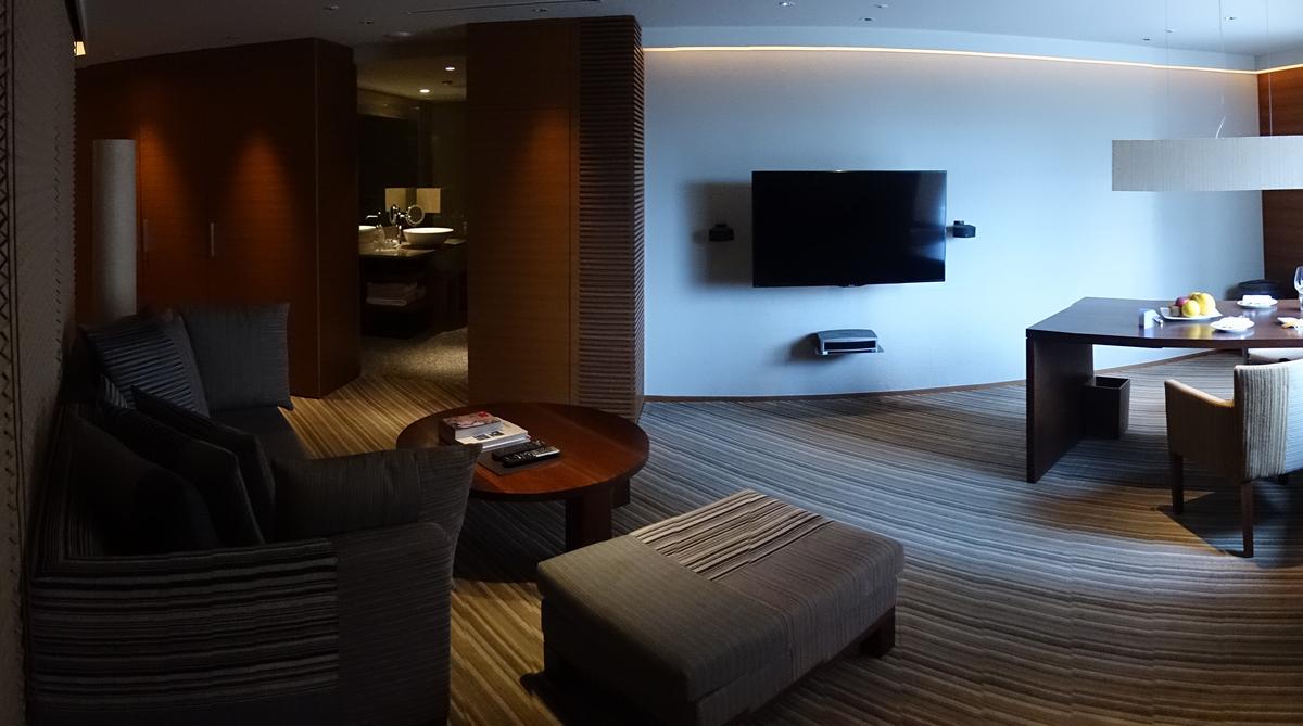 A contemporary hotel suite featuring a sofa, dining table, wall-mounted television, and an open doorway to a bathroom.