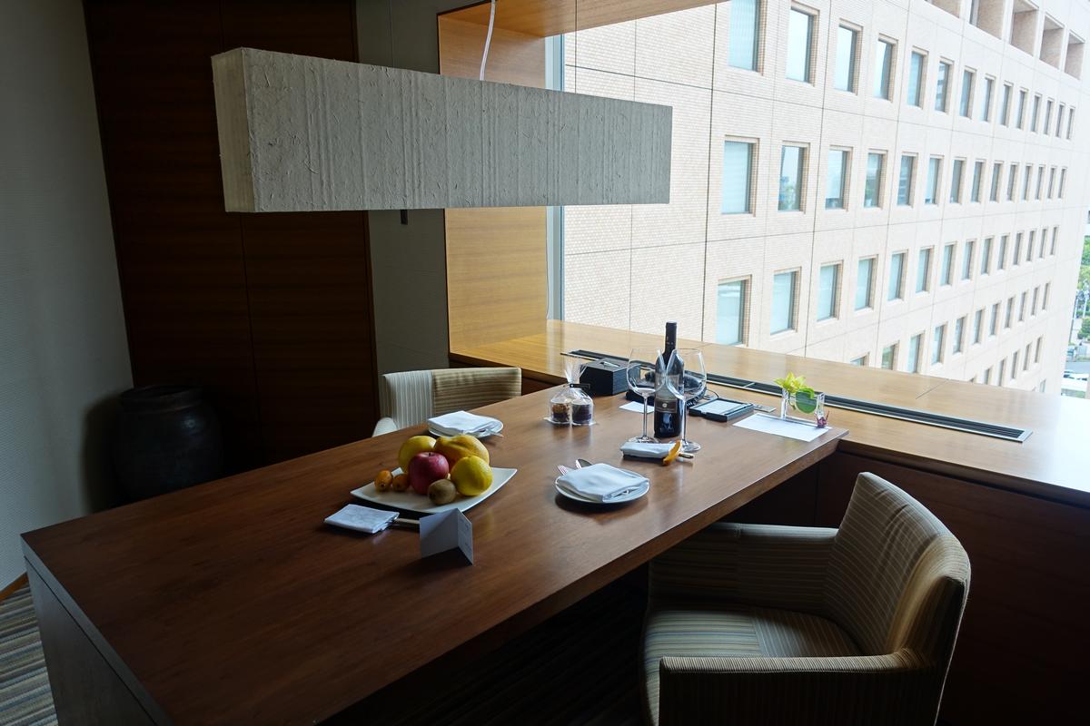 A long wooden table with a fruit bowl and wine in front of a large window with a city view.