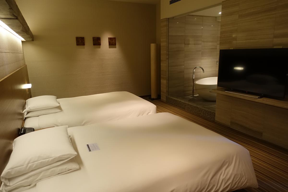 A hotel room with two twin beds, a television, and a glass-walled bathroom with a bathtub.