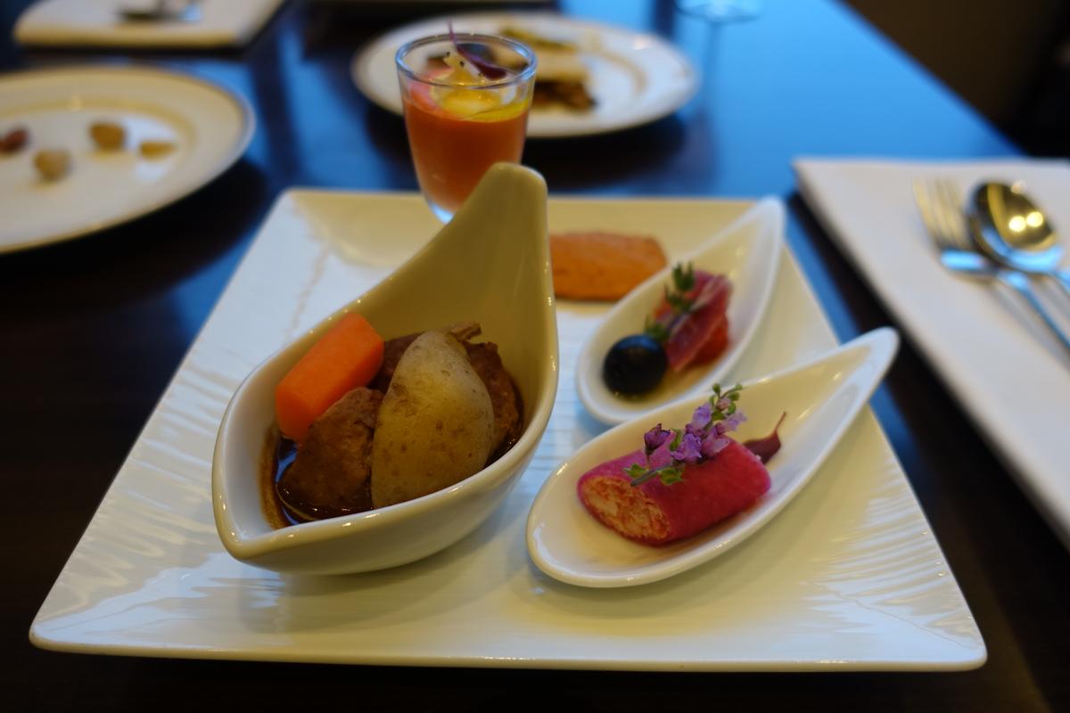An elegant white plate features a small bowl of stewed food, two spoon-shaped appetizers, and a glass of orange drink.
