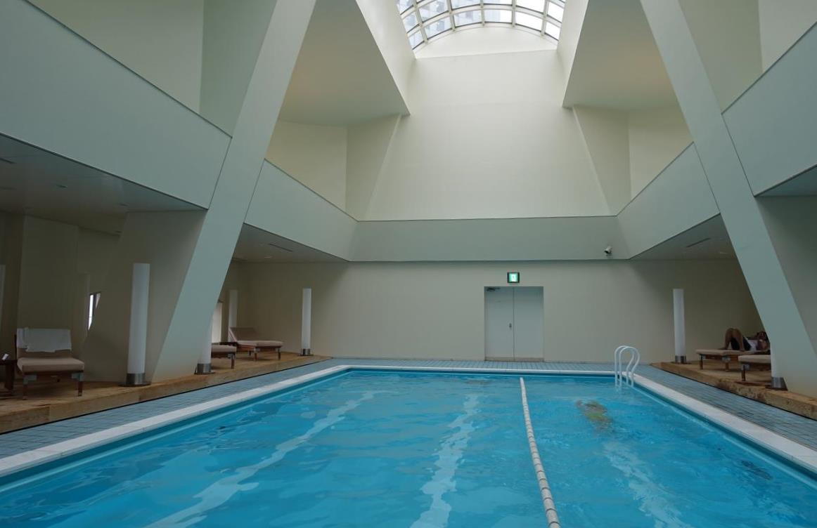 A modern indoor swimming pool at Hyatt Regency Tokyo, featuring a large skylight, lounge chairs, and a person swimming.