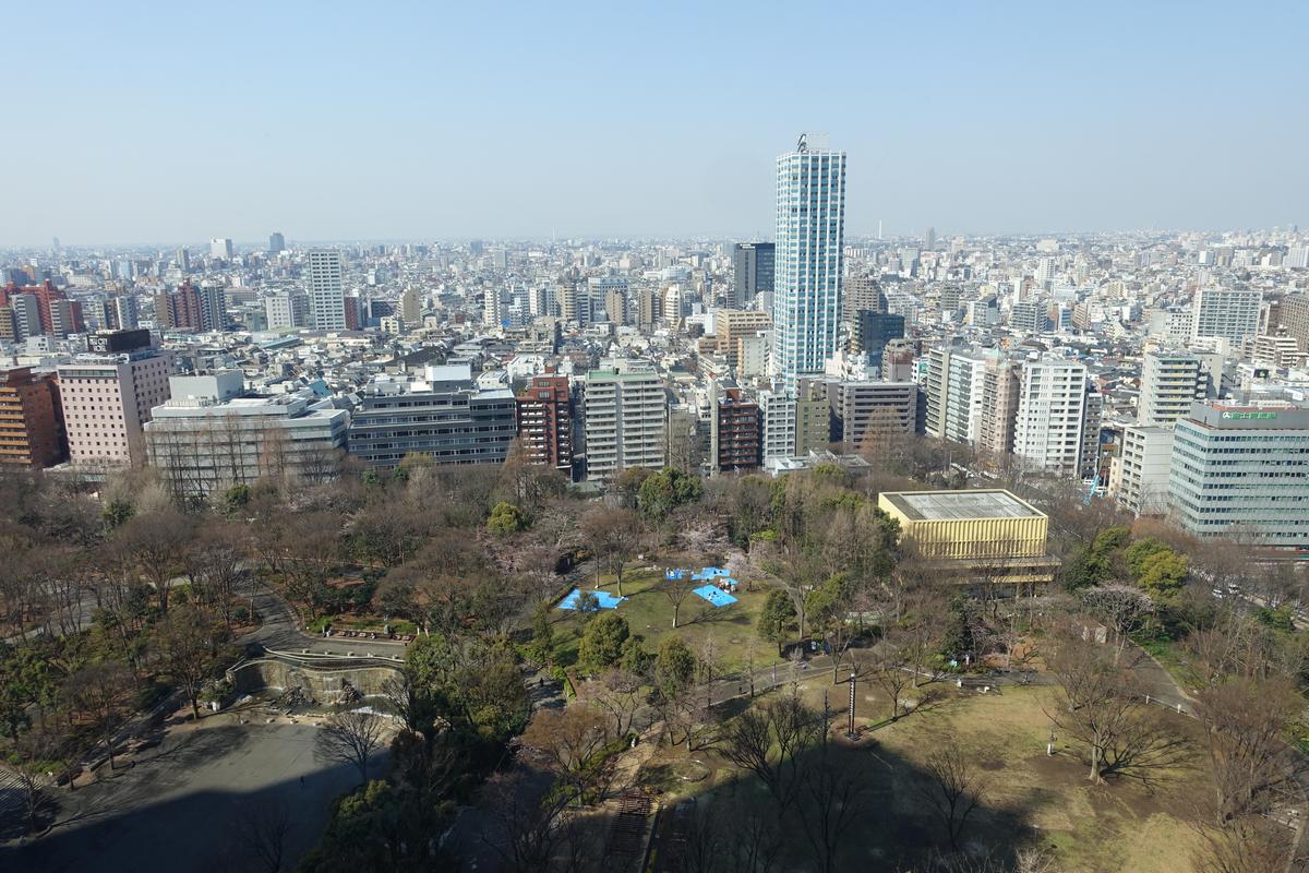 Wide aerial view of Tokyo showing a park below and a dense cityscape with numerous buildings and a tall skyscraper.