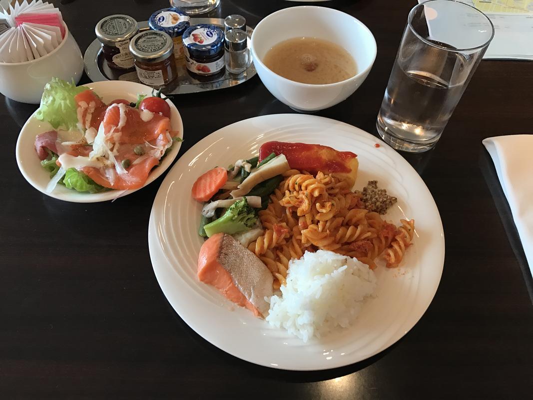 A hotel breakfast spread featuring a plate of salmon, rice, pasta, and vegetables, with a side salad, soup, and condiments.
