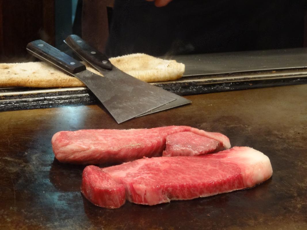 Raw Kobe beef steaks on a hot teppanyaki griddle with spatulas.