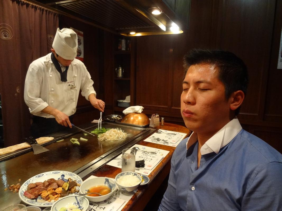 A chef cooks on a teppanyaki grill as a customer enjoys a meal of Kobe beef, eyes closed in appreciation.