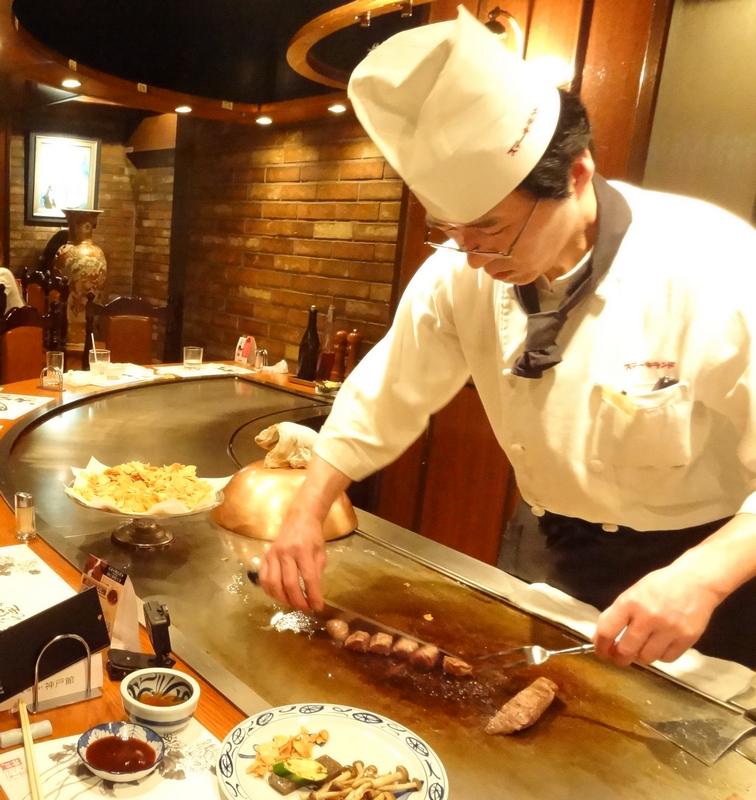 Chef in white uniform preparing Kobe beef on a large flat-top grill at a restaurant.