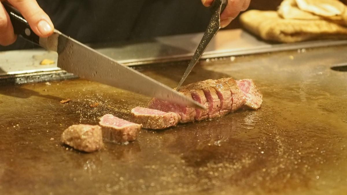 Hands slice pink Kobe beef on a sizzling teppanyaki griddle.
