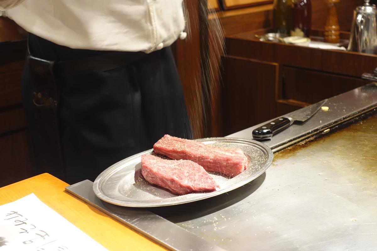 A chef seasons two raw cuts of Kobe beef on a plate next to a teppanyaki grill.