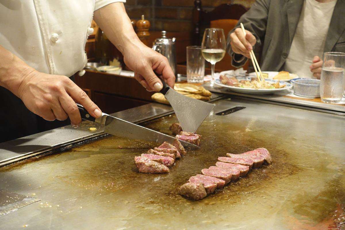 A chef slices cooked Kobe beef on a teppanyaki grill as a diner eats in the background.