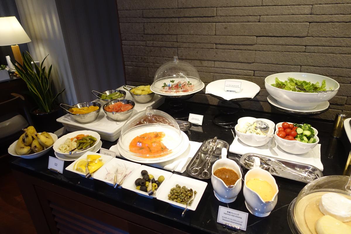 A diverse hotel buffet offers bowls of fresh fruit, salads, smoked salmon, and various dressings and toppings.