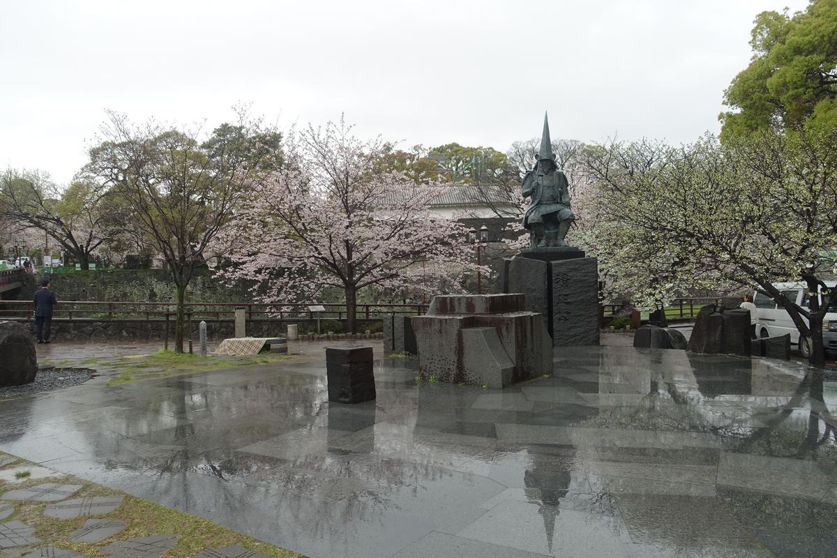 A bronze samurai statue stands amidst blooming cherry blossom trees on a wet, reflective pavement under an overcast sky.
