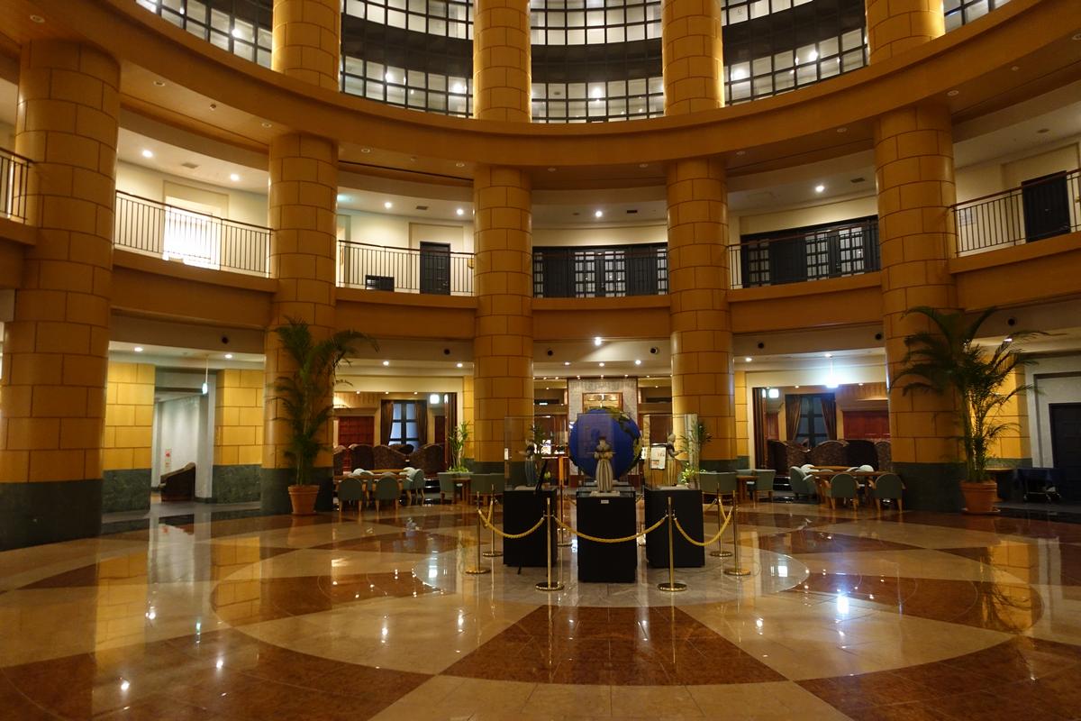 The grand multi-story lobby of the Hyatt Regency Fukuoka features numerous columns, a reflective tiled floor, and a large blue globe display.
