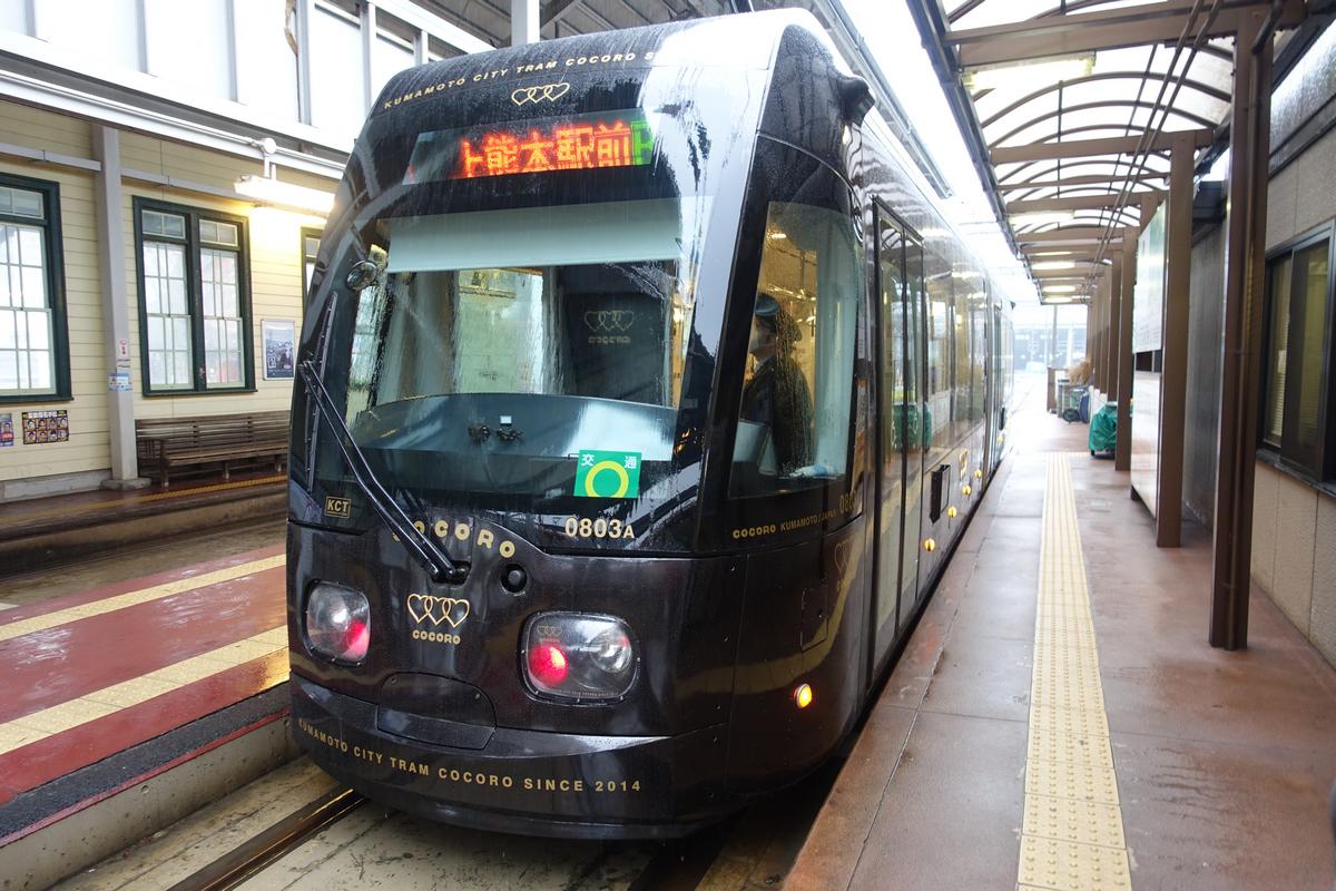 A sleek black Kumamoto City Tram Cocoro is stopped at a station platform on a wet day.