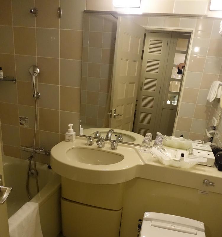 A beige-tiled hotel bathroom features a bathtub with a shower, a sink with toiletries, and a modern toilet.
