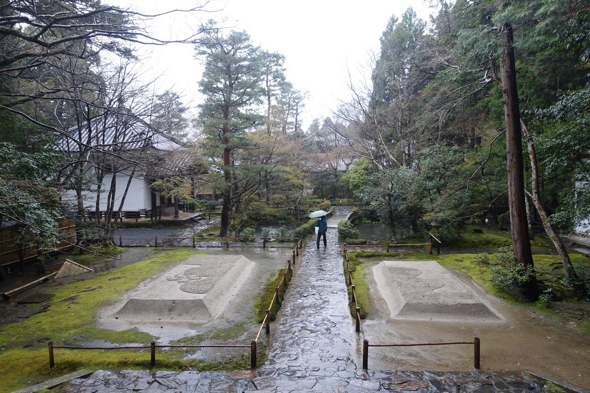 A person with an umbrella walks on a wet stone path between two raked sand gardens in a lush Japanese garden.