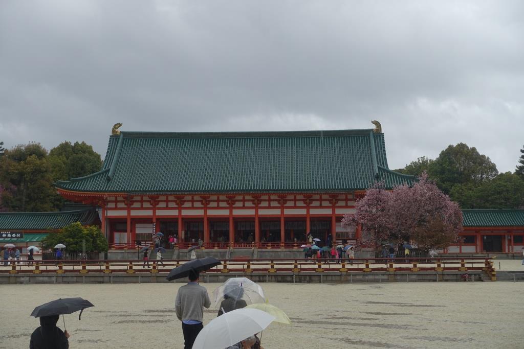 Heian Shrine on a rainy day, featuring a traditional green-roofed building, a pink cherry blossom tree, and visitors with umbrellas.