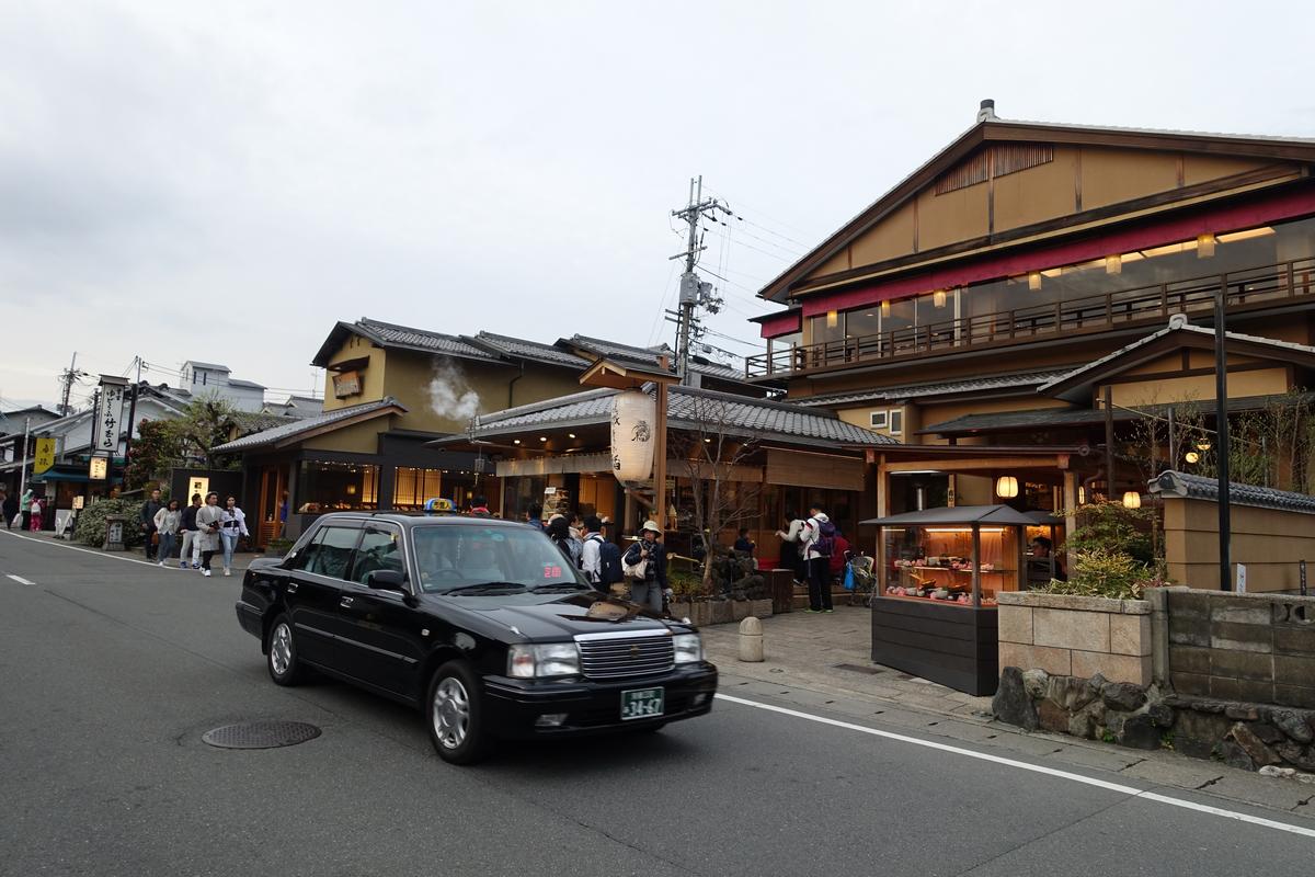 A black taxi drives down a street lined with traditional Japanese buildings and pedestrians under an overcast sky.