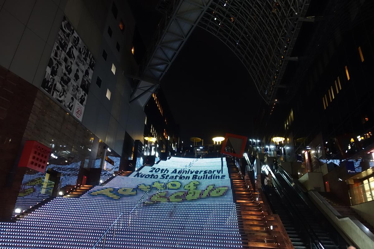 Large illuminated staircase at Kyoto Station building displaying a "20th Anniversary" message at night.