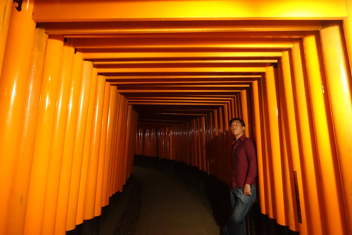 A person stands within a vibrant orange torii gate tunnel at Fushimi Inari Shrine.
