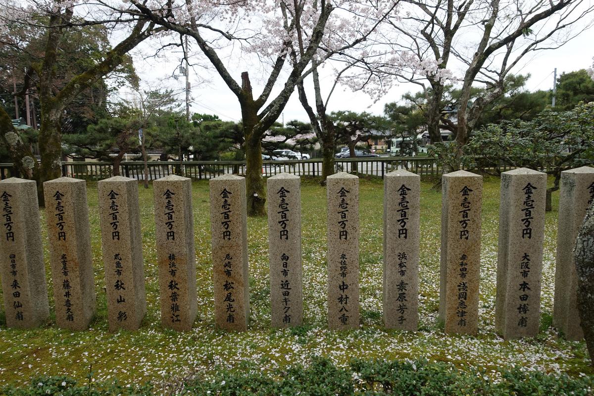 Stone pillars with Japanese writing stand among fallen cherry blossoms and flowering trees.