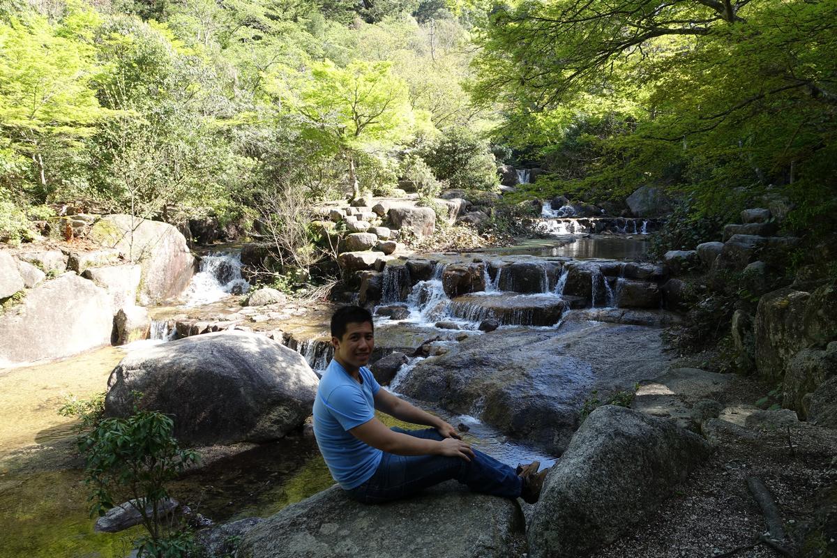 A smiling man rests on a large rock next to a cascading stream in a lush forest on Miyajima Island.