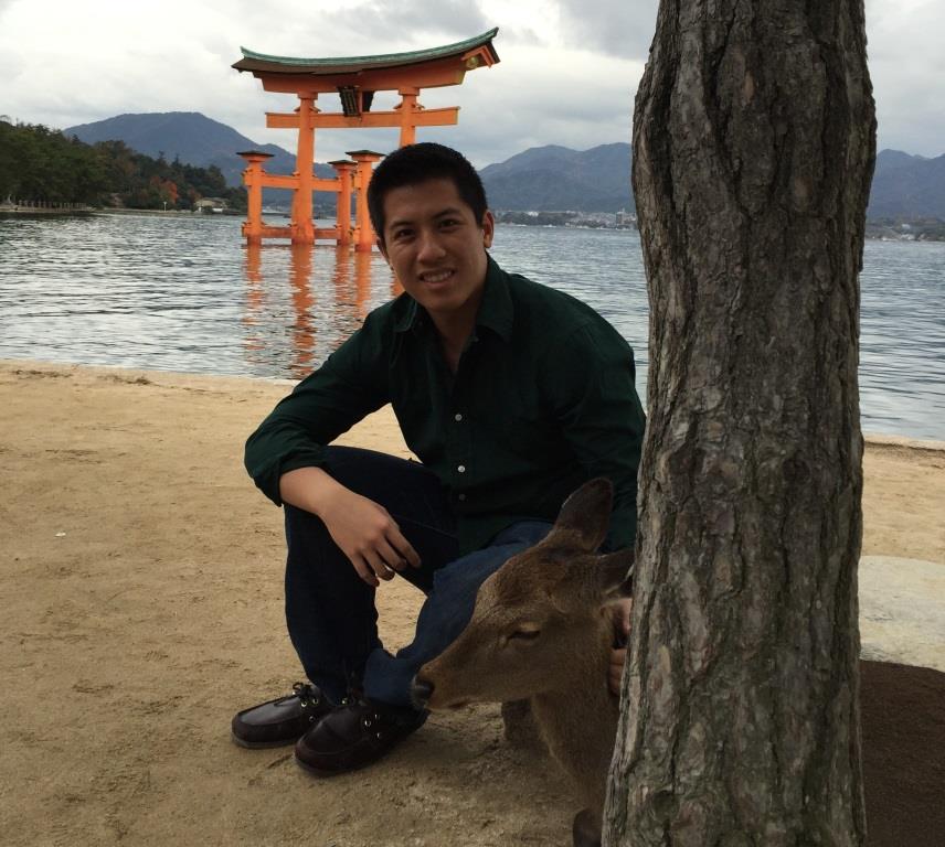 A man kneels on a sandy beach next to a deer, with Miyajima's iconic orange torii gate visible in the water behind him.
