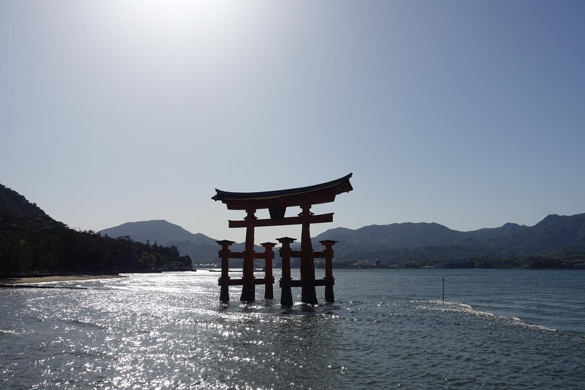 The iconic floating torii gate of Miyajima Island stands in the water against a bright sky and distant mountains.