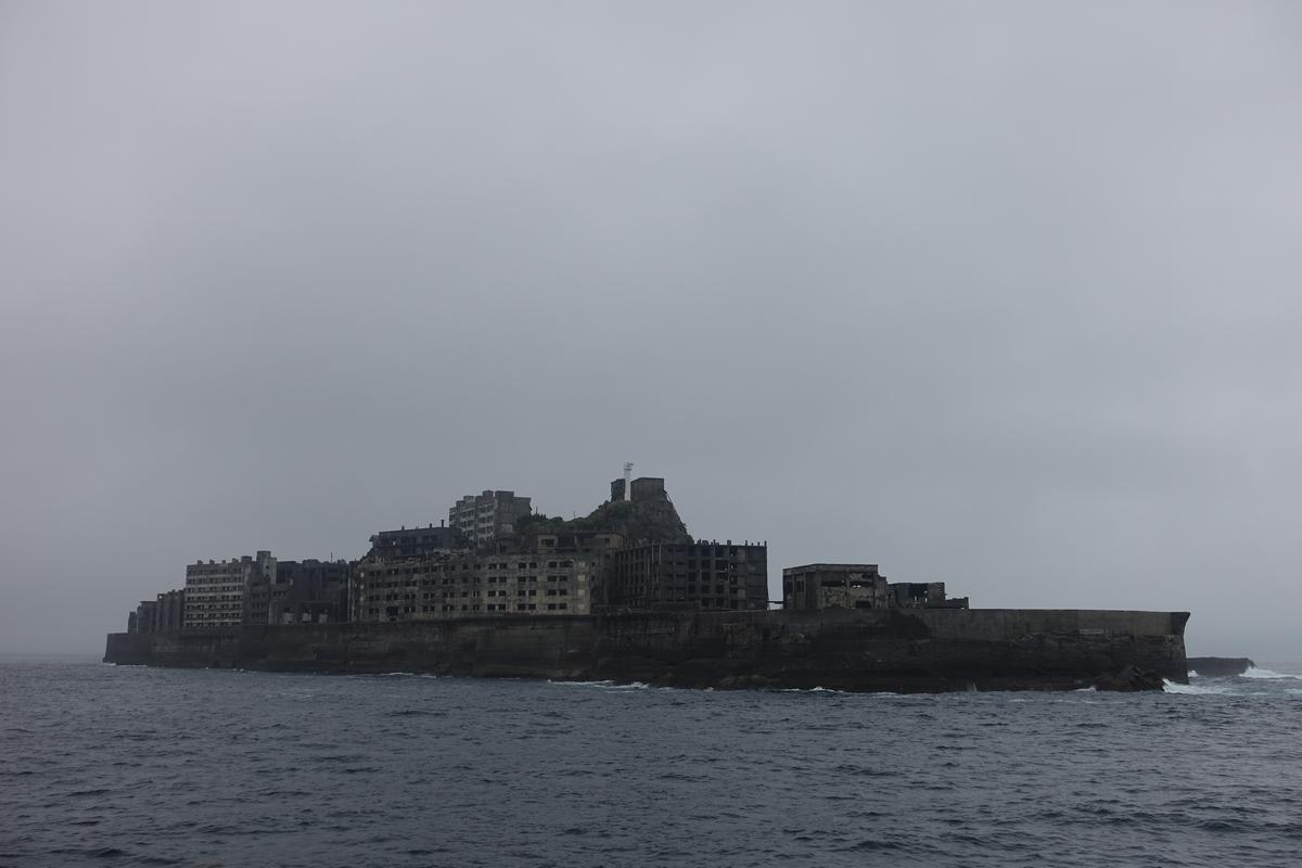 Crumbling buildings of Battleship Island (Hashima) rise from the dark ocean under a grey sky.