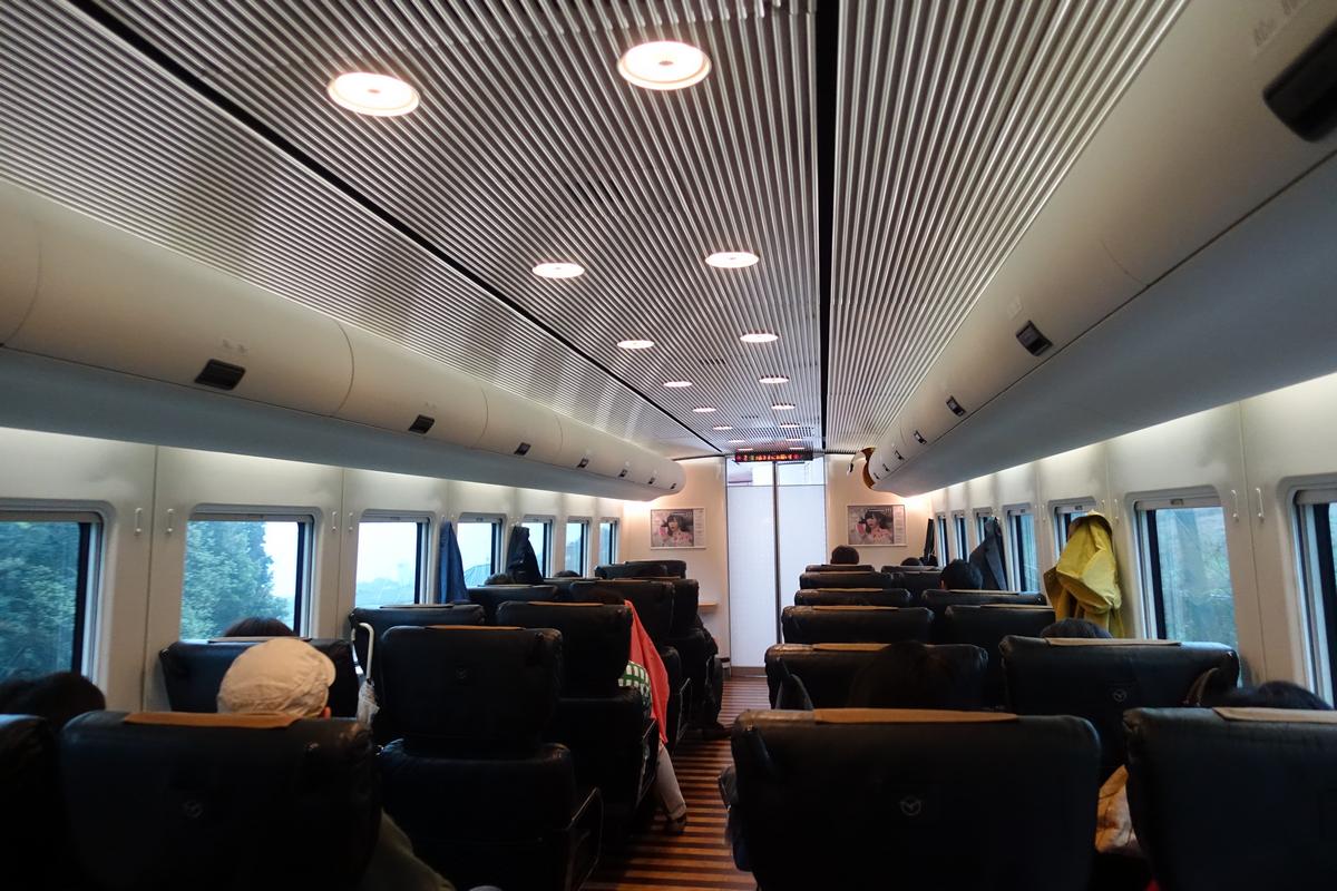 Interior view of a train carriage with rows of black seats, some occupied by passengers, and windows showing trees outside.