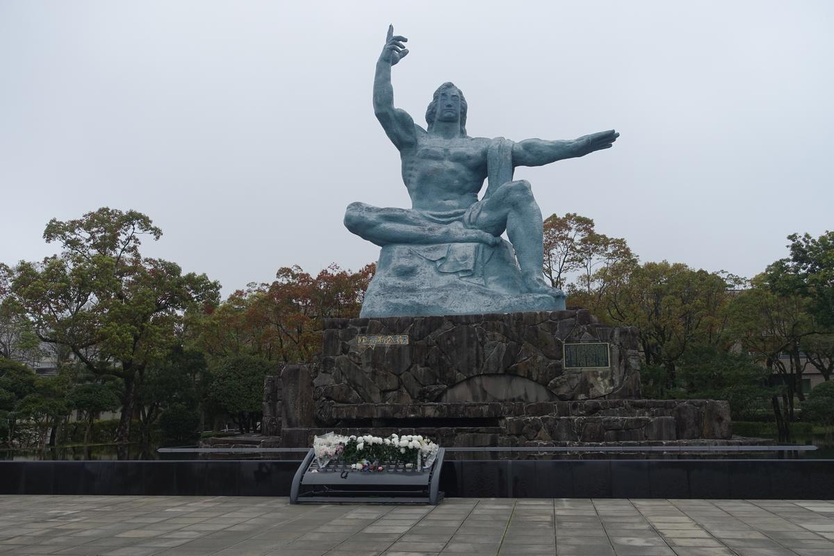 The blue-grey Nagasaki Peace Statue, a seated muscular man with one arm raised and one extended, at Nagasaki Peace Park with flowers at its base.
