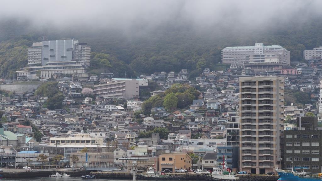 A dense Nagasaki cityscape rises from a waterfront up a steep, fog-covered hill.