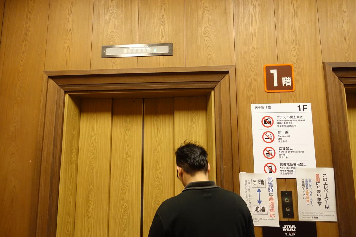 A person stands facing a wooden elevator door on the first floor, next to informational signs in Japanese and English.