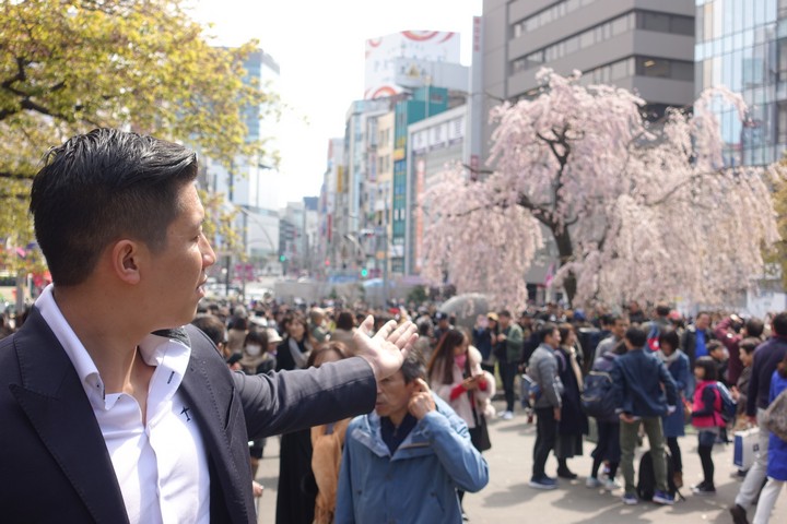 A man gestures on a crowded city street with blooming pink cherry blossom trees and buildings in the background.