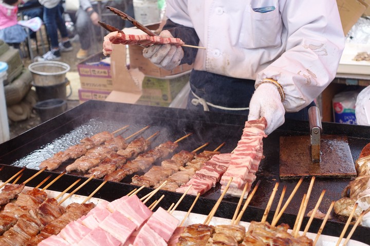 A street food vendor cooks various meat skewers on a large griddle.