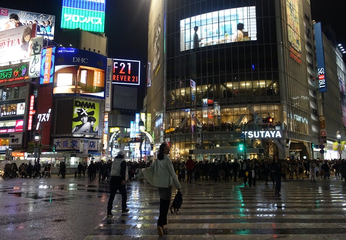 A person walks across a wet city intersection at night, surrounded by brightly lit buildings and neon signs.