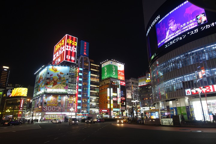 A bustling Shibuya street scene at night, illuminated by colorful neon signs and large digital billboards.