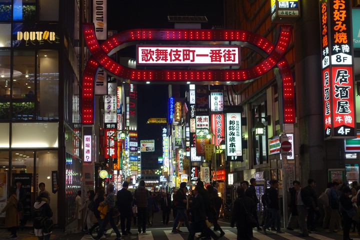 A busy street in Tokyo's Kabukicho district at night, illuminated by a large red archway and numerous neon signs, with people crossing.