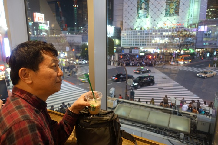 A man holds a Starbucks drink, looking out a window at the illuminated Shibuya Crossing at night.