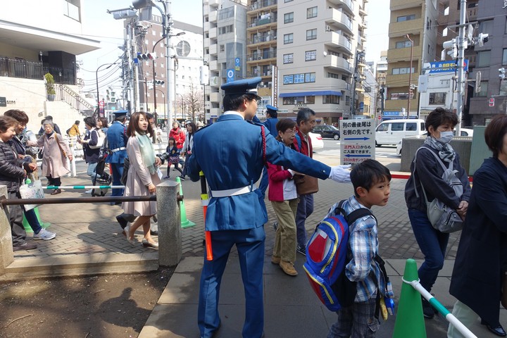 A uniformed Japanese police officer directs pedestrians on a sunny city street.