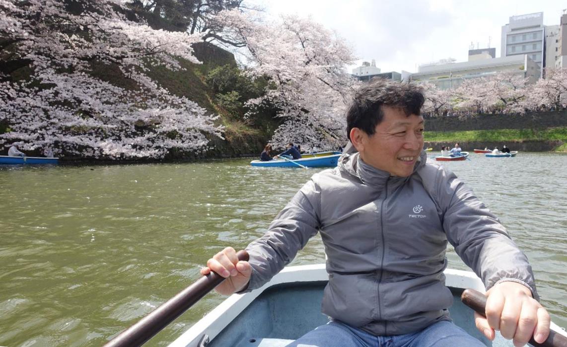 A man smiles while rowing a boat on the Chidorigafuchi moat, surrounded by blooming cherry blossom trees and other boats.