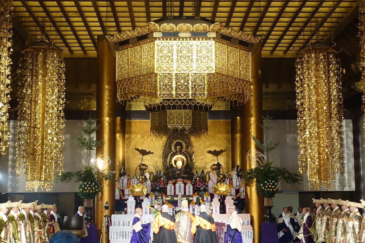 A golden-ornamented temple hall with a Buddha statue at the altar and robed figures participating in a ceremony.