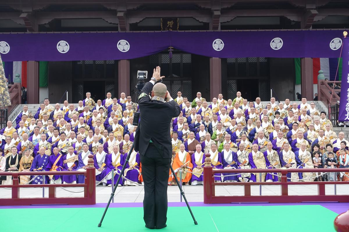 A photographer takes a group photo of people in traditional robes seated on temple steps under a large purple banner.