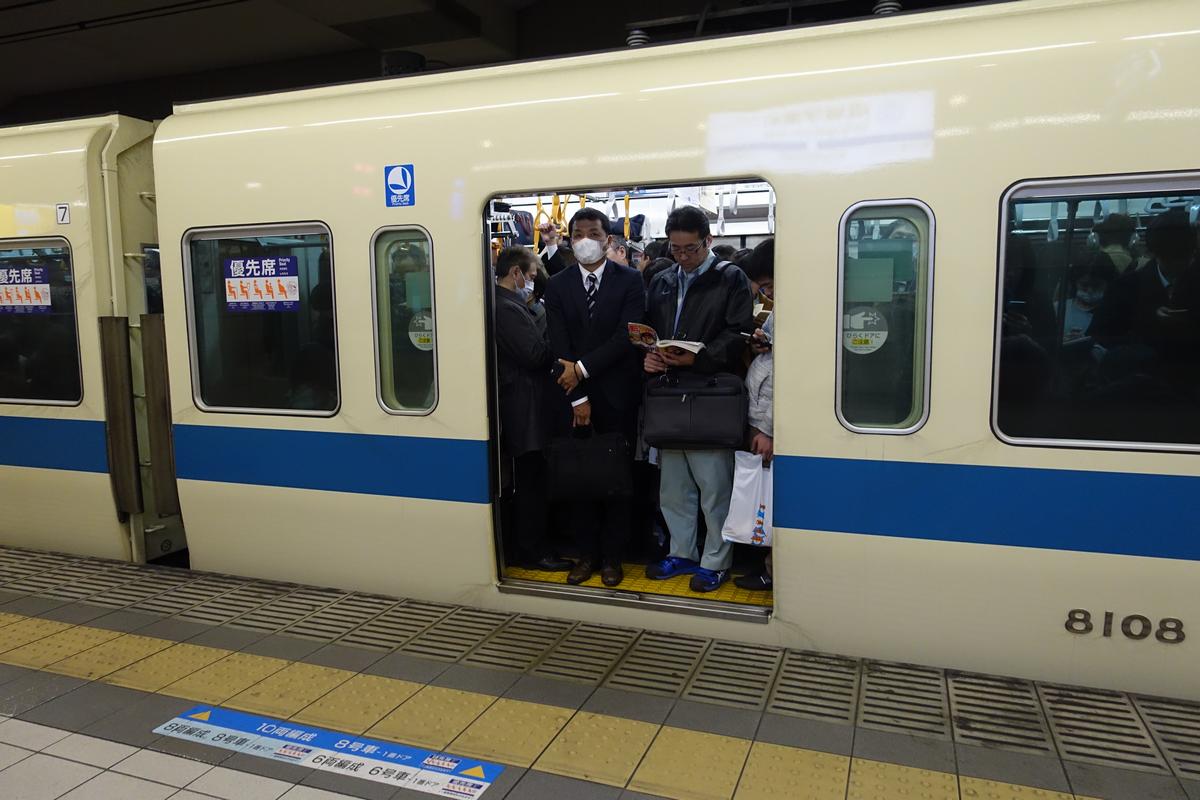 A crowded train with open doors stands at a station platform, showing passengers inside including a man in a face mask in the doorway.